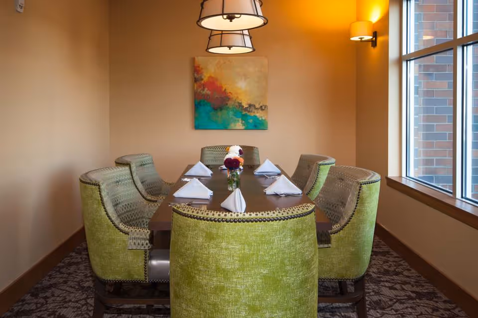Small dining room with a wooden table set for six, green upholstered chairs, folded napkins, a floral centerpiece, pendant lights, artwork on the wall, and a window to the right.