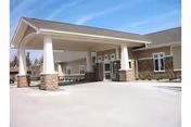 Entrance of a single-story senior living facility building with a covered drop-off area supported by white columns with stone bases, beige siding, and multiple windows under a clear blue sky.