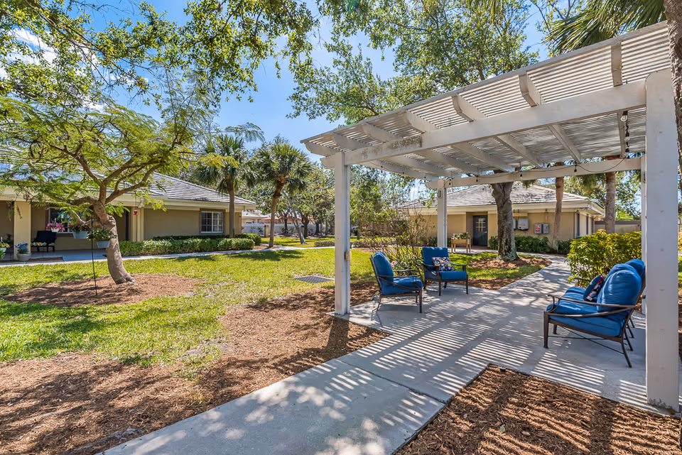 Outdoor seating area with blue cushioned chairs under a white pergola surrounded by trees and greenery, with single-story buildings in the background under a clear blue sky.