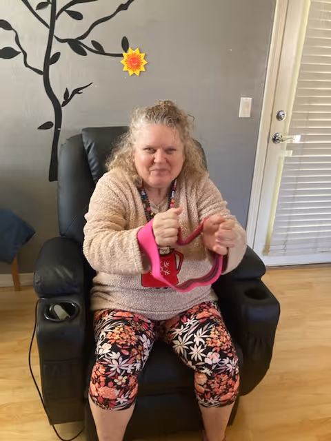 A woman sits in a black recliner in a living room holding a pink exercise band.