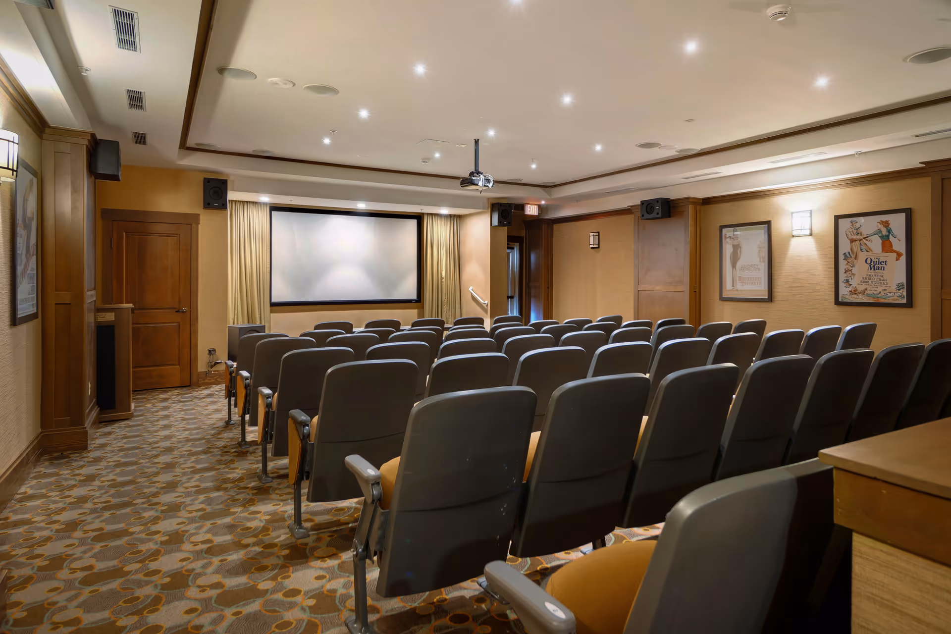 A small theater room with rows of gray cushioned seats facing a large white projection screen. The room has beige walls with wooden trim, framed movie posters on the walls, recessed ceiling lights, and a ceiling-mounted projector.