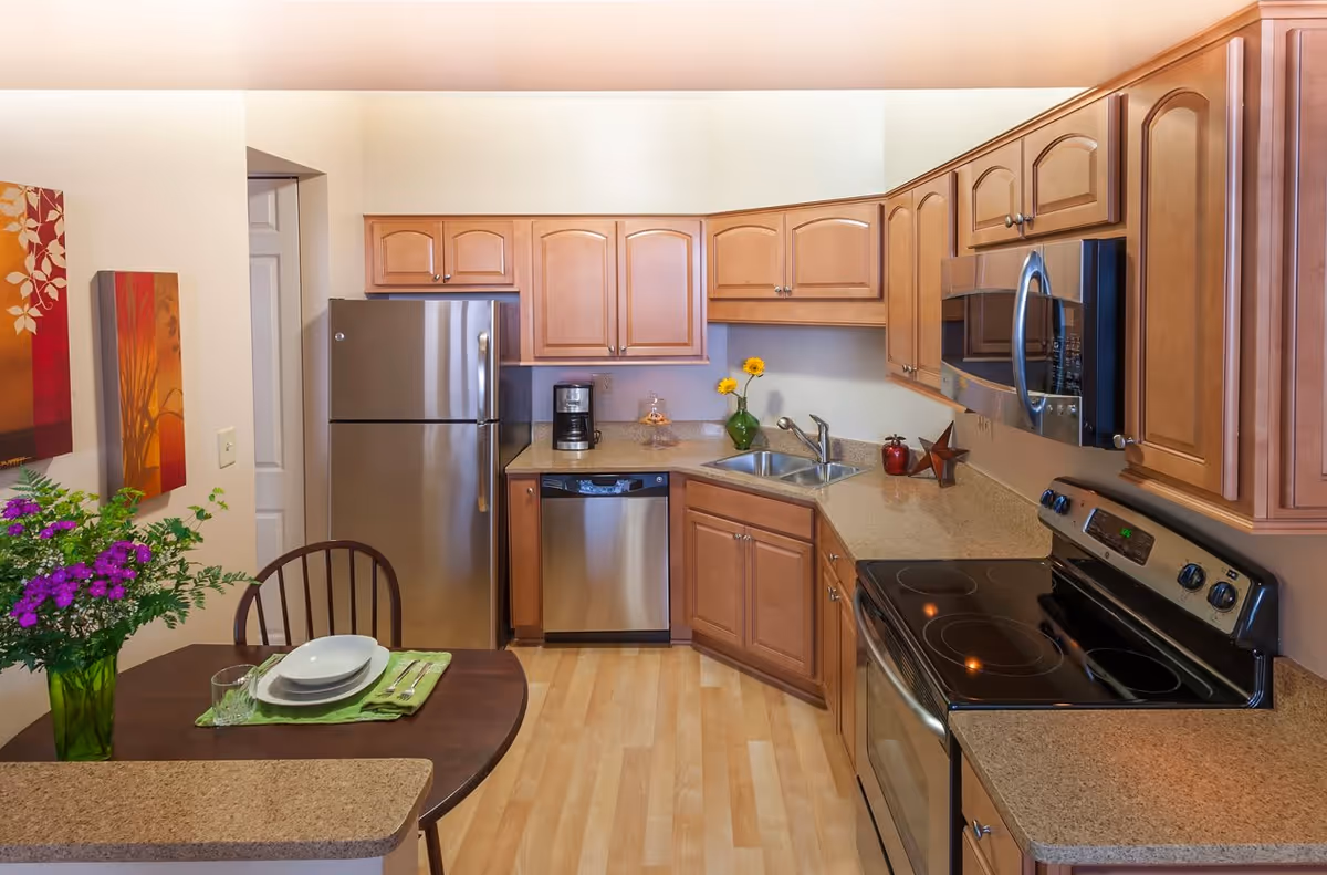 A modern kitchen with light wooden cabinets, stainless steel appliances including a refrigerator, dishwasher, microwave, and electric stove. There is a small dining table with a single place setting and a vase of purple flowers. The kitchen has light wood flooring and decorative wall art.