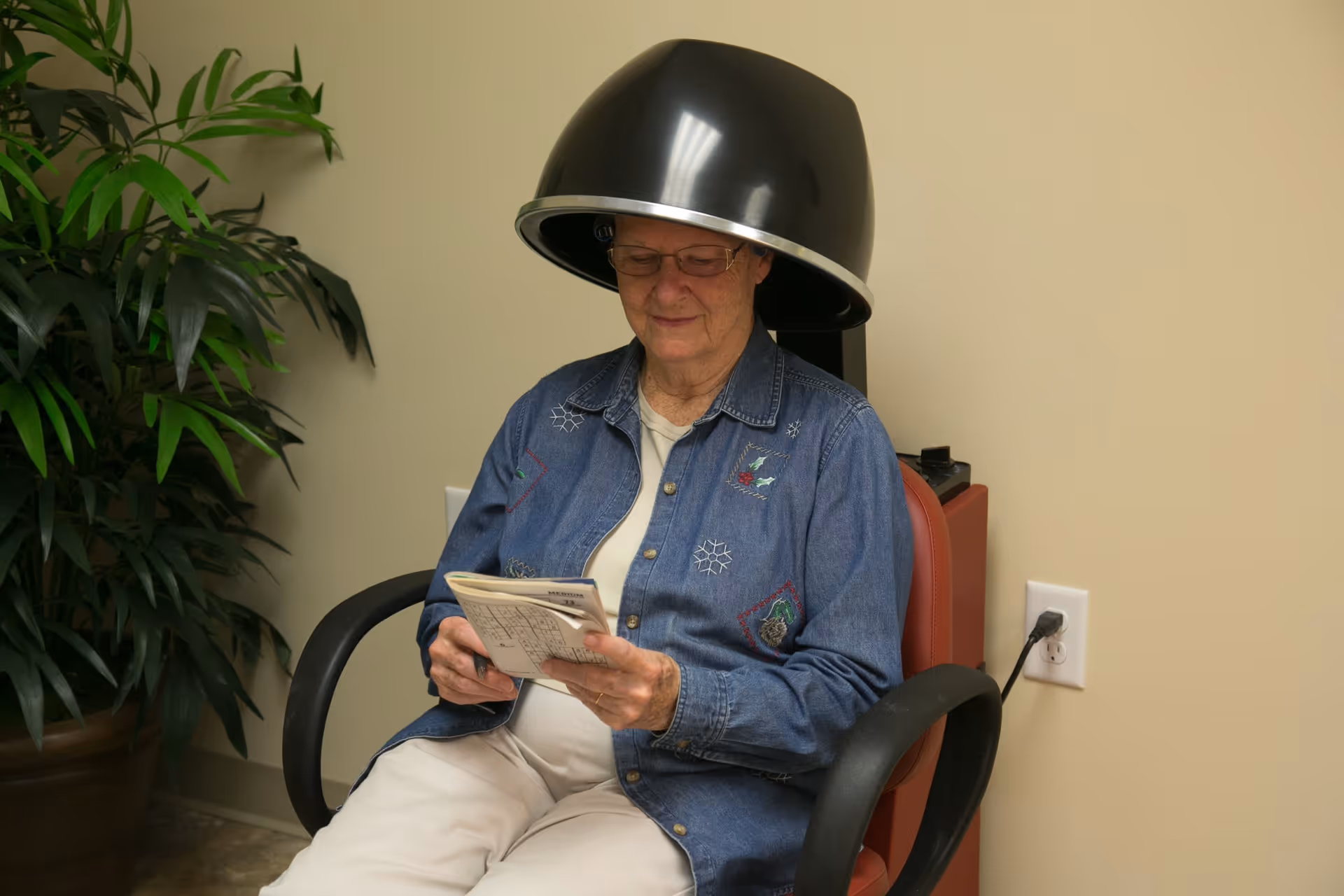 An elderly woman wearing glasses sits under a black hair dryer in a salon chair, reading a newspaper. She is dressed in a blue denim shirt and light-colored pants. There is a large green potted plant to her left and a beige wall with an electrical outlet behind her.