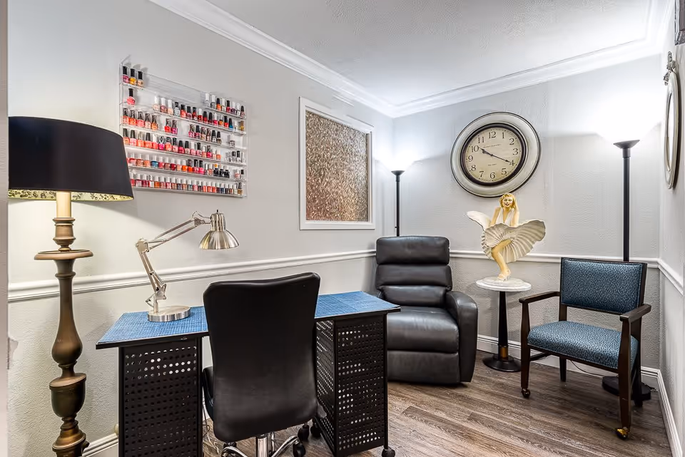 Interior salon-style room with a manicure desk, nail polish display, recliner, chairs, lamps, and a wall clock.