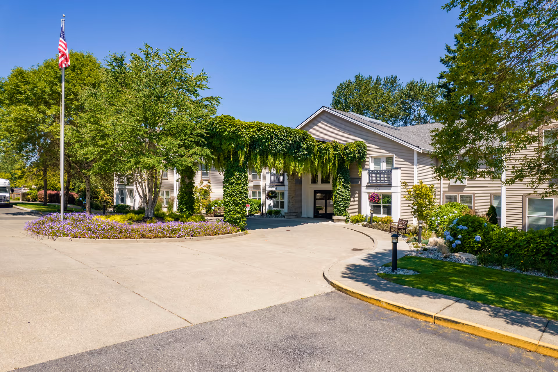Exterior view of GenCare Lifestyle Federal Way facility on a sunny day, showing a two-story building with beige siding, a driveway with a circular flower bed featuring purple flowers and an American flag, and an entrance covered with green ivy. Trees and shrubs surround the building.