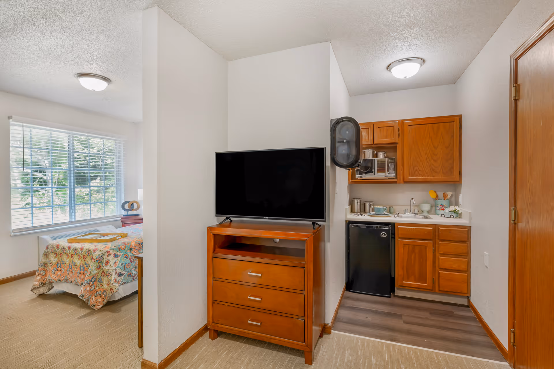 Interior view of a senior living facility room showing a small kitchenette with wooden cabinets, a mini fridge, microwave, and sink. Adjacent to the kitchenette is a wooden dresser with a flat-screen TV on top. In the background, there is a bedroom area with a bed covered in a colorful patterned quilt and a large window with blinds letting in natural light.