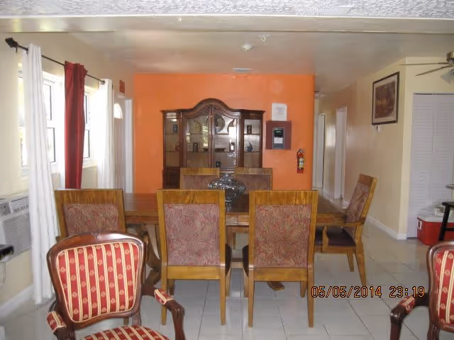 Interior view of a dining room in an assisted living facility featuring a wooden dining table with six upholstered chairs. A wooden china cabinet is against an orange accent wall in the background. The room has white tiled flooring, cream-colored walls, windows with white and red curtains, and an air conditioning unit on the left wall.