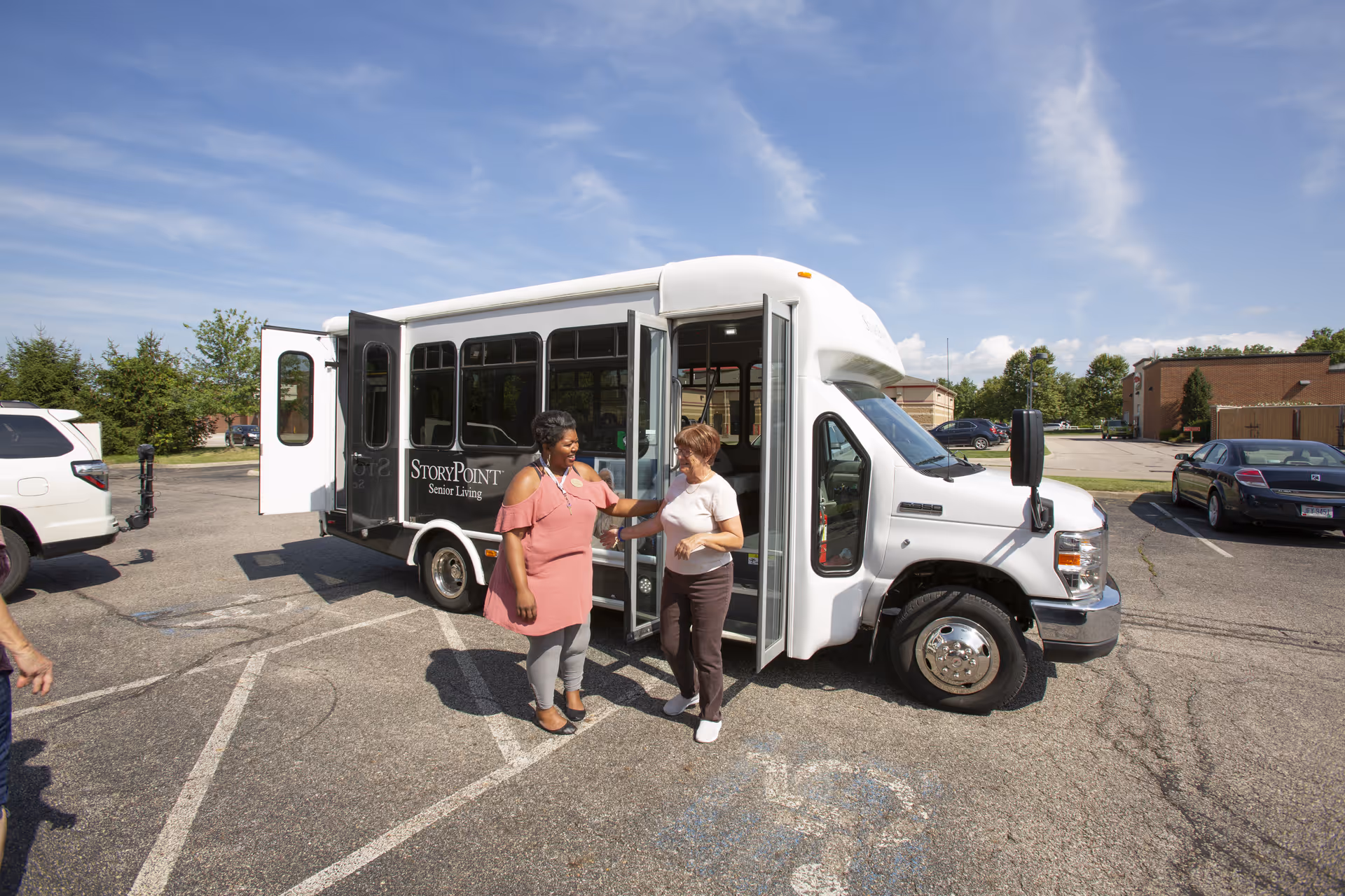 Two women standing outside a white StoryPoint Senior Living shuttle bus in a parking lot on a sunny day with a blue sky and some clouds.