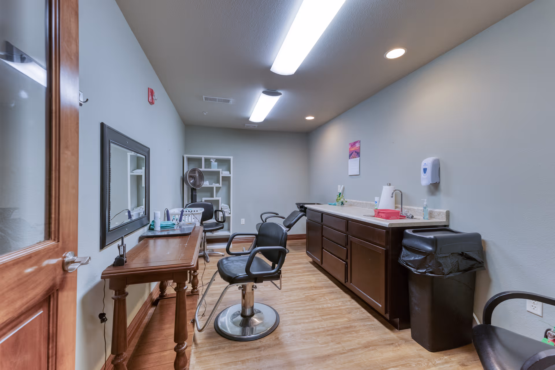 Interior view of a small salon or grooming room with a wooden table, a large mirror on the wall, two black salon chairs, a hair dryer, a countertop with cabinets underneath, a trash bin, and various grooming supplies.