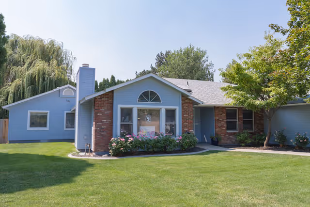 Single-story residential building with blue siding and red brick accents, surrounded by green grass, bushes, and trees under a clear sky.