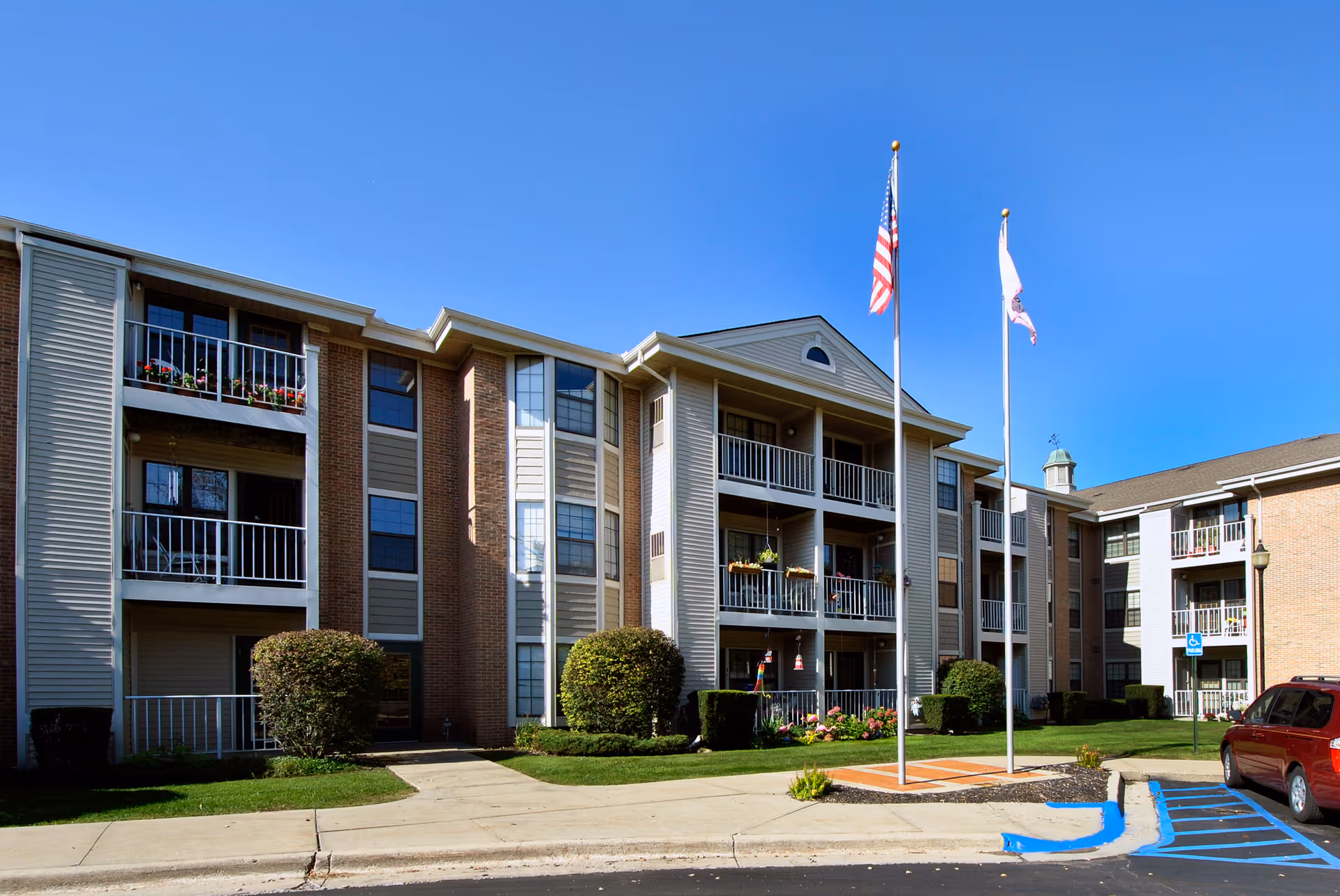 Exterior view of a three-story senior living facility building with balconies, two flagpoles with the American flag and another flag, well-maintained landscaping, and a parking area with a handicap parking space.