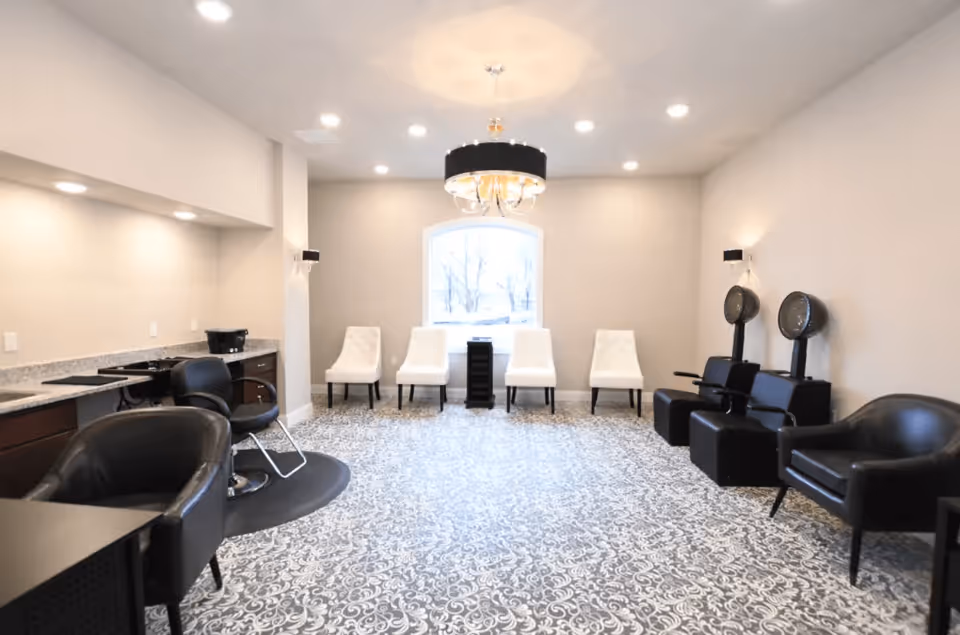 A bright and spacious salon area with patterned carpet flooring, a row of four white chairs against the far wall under a large window, two black salon hair dryers on the right, and black salon chairs on the left near a countertop with cabinets and a sink. A modern chandelier hangs from the ceiling.