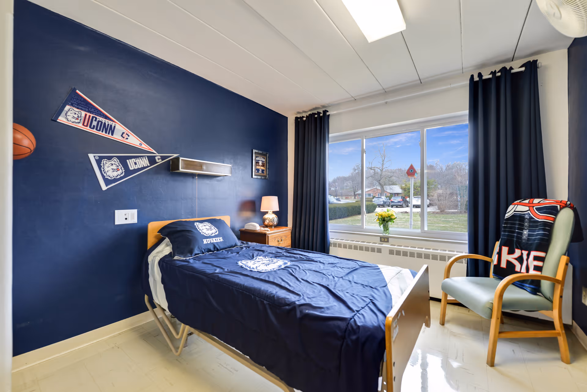 A navy-decorated single bedroom with UConn bedding, a nightstand and lamp, a large window with curtains, and a chair with a blanket.