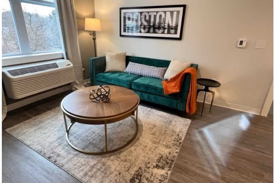 Living room with a green velvet sofa, round wooden coffee table on a patterned rug, floor lamp, and a window air conditioner.