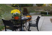 Outdoor patio with a round table and chairs, a potted yellow flower on the table, and stone landscaping in the background.