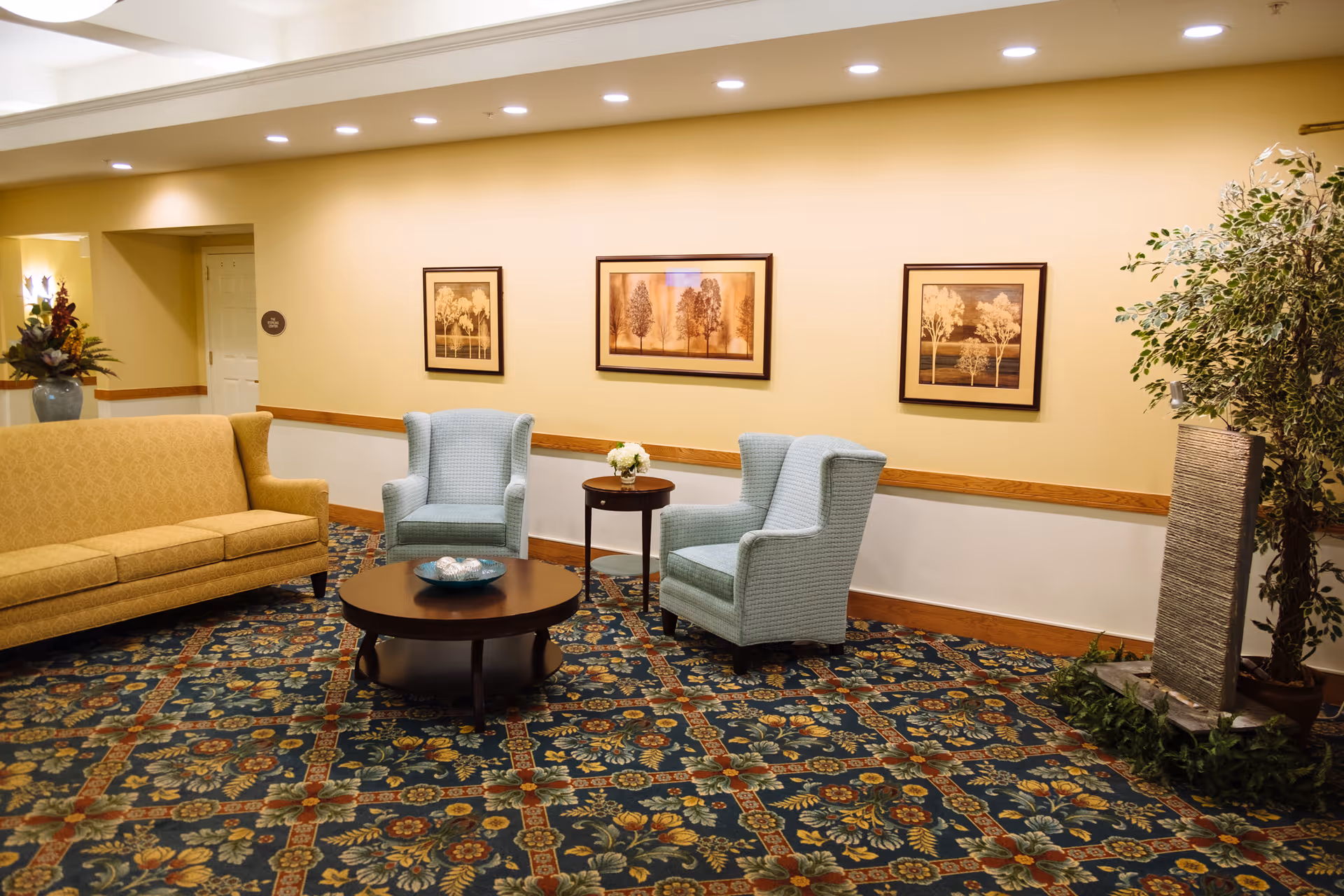 A cozy seating area in a senior living facility with a yellow sofa, two light blue armchairs, a round wooden coffee table, and a small side table with a flower arrangement. The walls are painted beige and decorated with three framed pictures of trees. The floor is covered with a patterned carpet, and there is a tall plant and a decorative water feature in the corner.