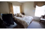 A small bedroom with a bed covered in a floral bedspread, two windows with curtains, a television on a stand, and a wooden chair near the window.