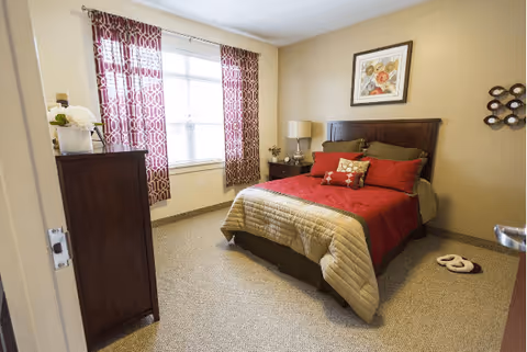 A cozy bedroom with a double bed dressed in red and beige bedding, a wooden headboard, and decorative pillows. The room features beige walls, a window with patterned red and white curtains, a wooden dresser with a flower vase, a bedside table with a lamp and small plant, a framed artwork above the bed, and a pair of slippers on the carpeted floor.