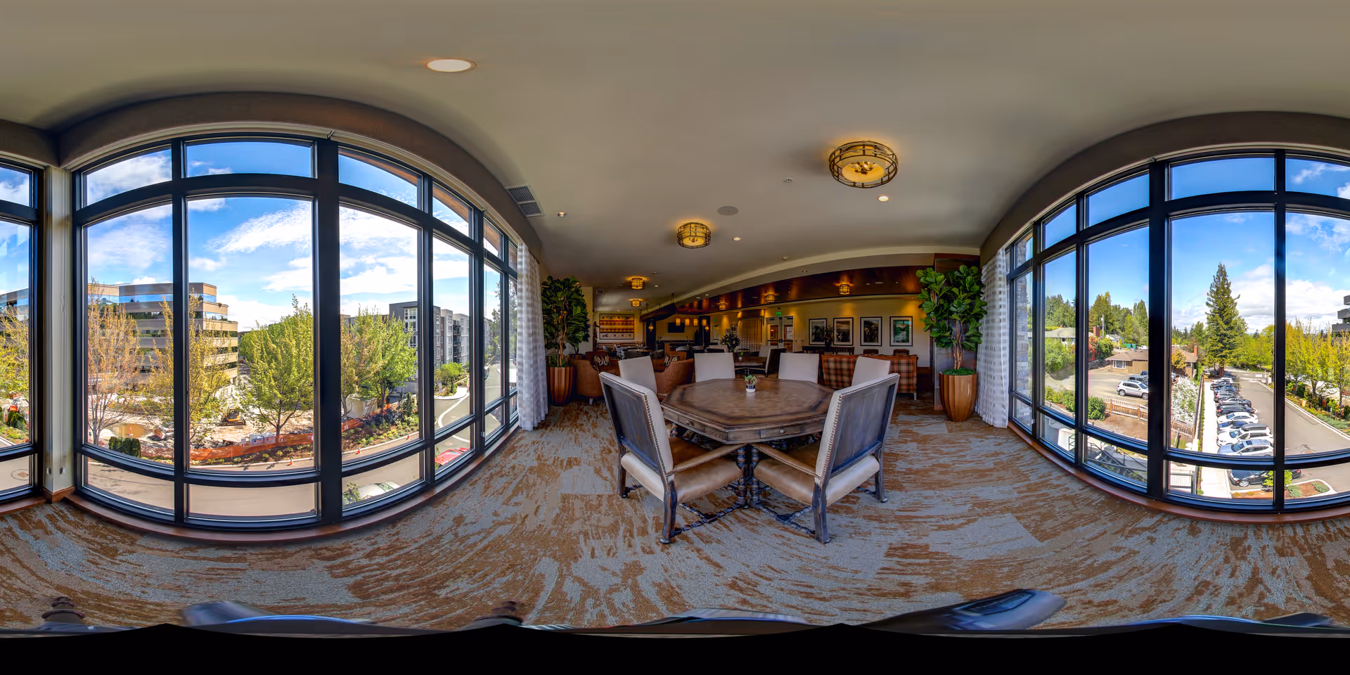 A spacious and well-lit common area with large curved windows offering a panoramic view of trees, buildings, and a parking lot outside. Inside, there is a round table with six chairs in the center, carpeted flooring, potted plants, and framed artwork on the walls. The ceiling has modern light fixtures.