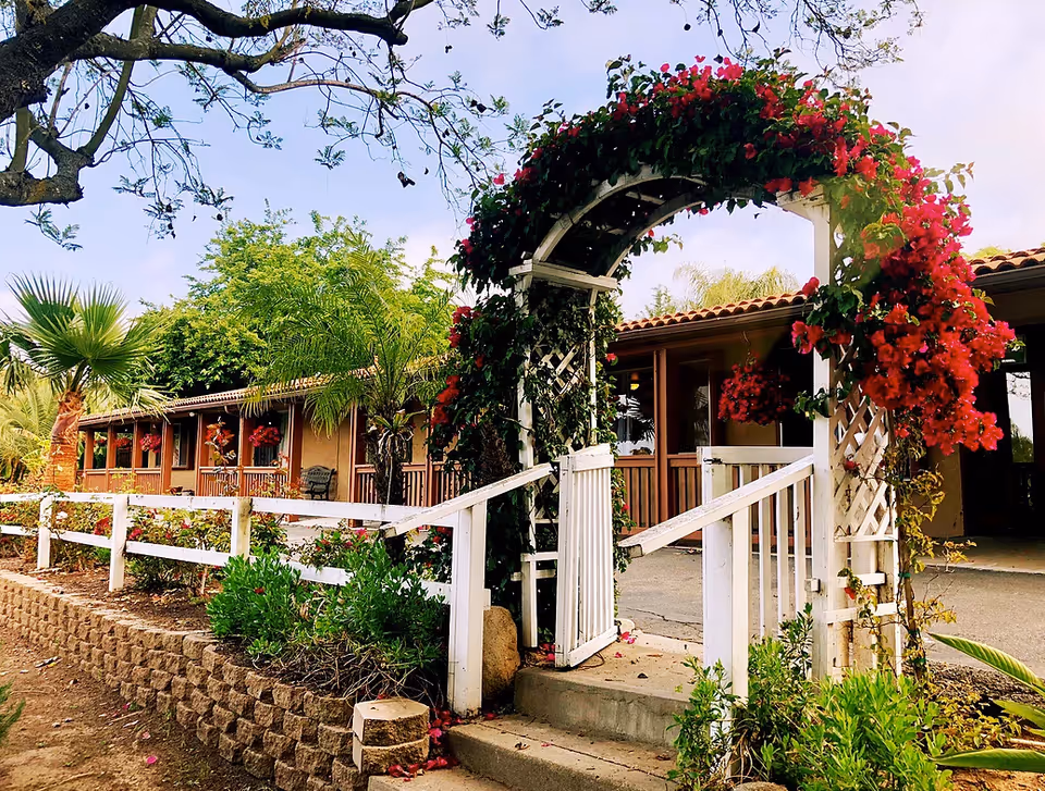 Entrance to a senior living facility with a white wooden arch covered in vibrant red flowers. The pathway leads to a building with a tiled roof and hanging flower baskets. Surrounding the area are various green plants and trees under a partly cloudy sky.