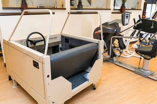 Indoor exercise room with a driving simulator machine in the foreground and various fitness equipment including a rowing machine and stationary bike in the background. The room has wooden flooring and mirrors on the walls.
