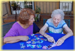 Two elderly women sitting at a table covered with a blue tablecloth, engaging in assembling a jigsaw puzzle together. They are smiling and appear to be enjoying the activity. The setting appears to be an outdoor patio area with railings and some plants in the background.
