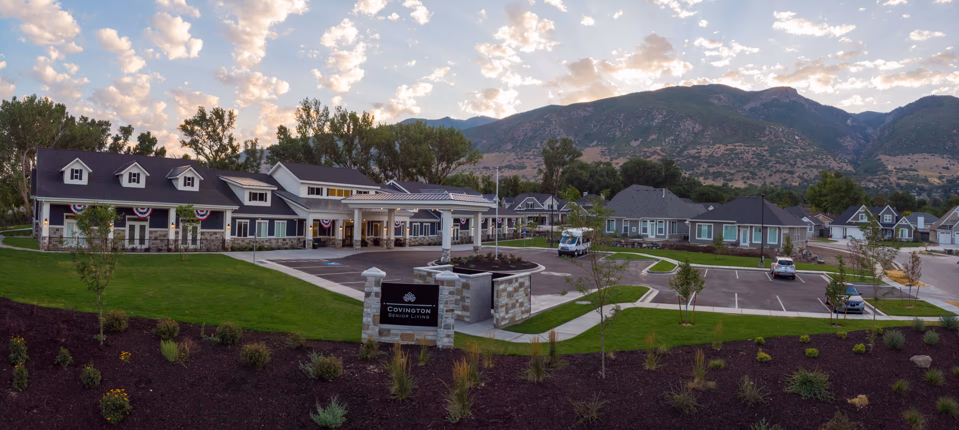 Wide exterior view of Covington Senior Living of Farmington facility with multiple buildings, parking lot, landscaped green lawns, and mountains in the background under a partly cloudy sky.