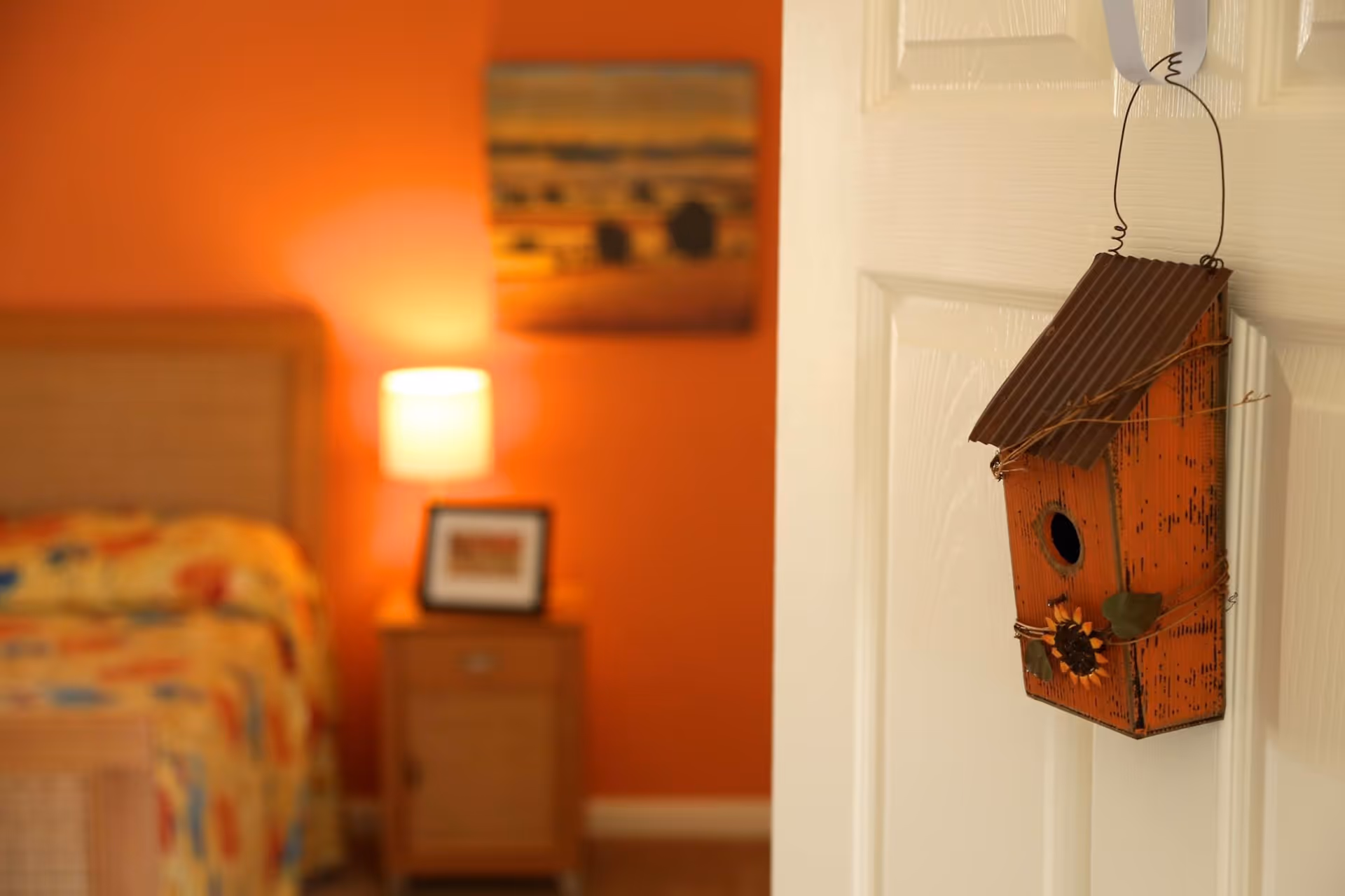 A decorative orange birdhouse hangs on a white door with a blurred orange-walled bedroom, bed, nightstand and lamp visible in the background.