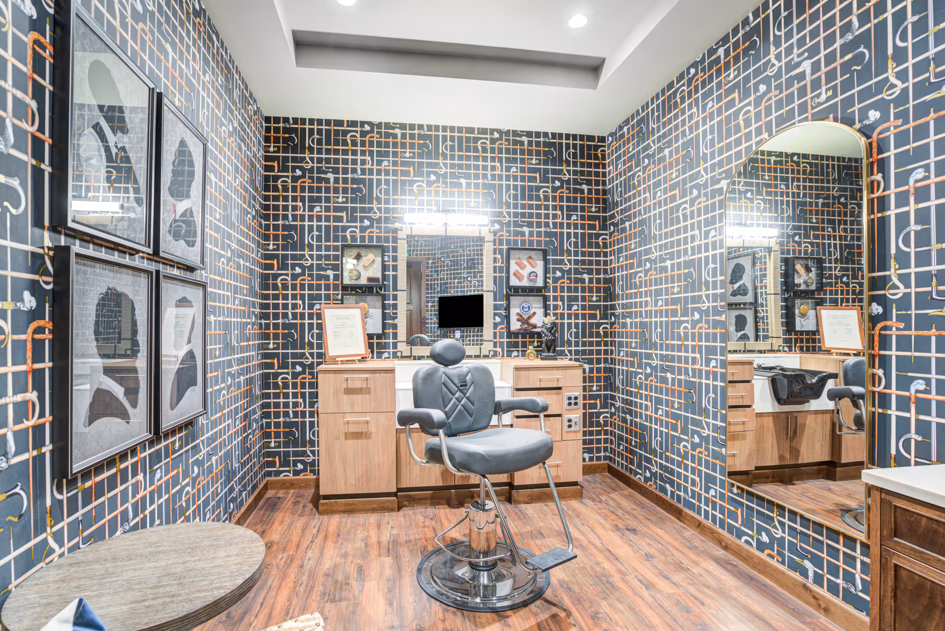 A stylish salon room with a black and orange patterned wallpaper, a large mirror on the right wall, a black salon chair in the center, wooden cabinets with a countertop and a sink, and framed artwork on the left wall. The floor is wooden and the room is well-lit with ceiling lights.
