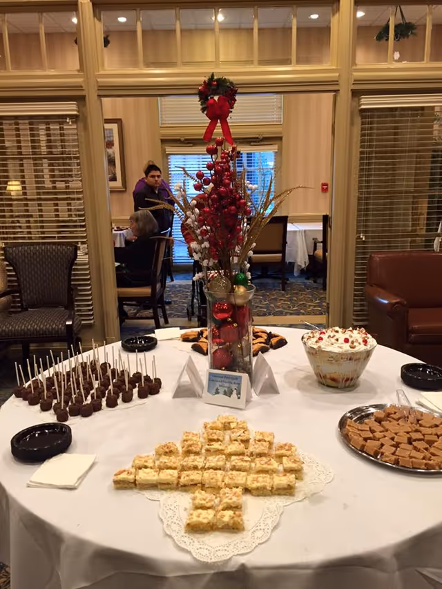 A round table covered with a white tablecloth displaying various desserts including cake pops, layered dessert in a glass bowl, fudge squares, and small square cake pieces arranged in a cross shape. A tall glass vase with red and gold holiday decorations is in the center of the table. In the background, there are chairs, a person sitting, and a person standing in a room with beige walls and windows with blinds.