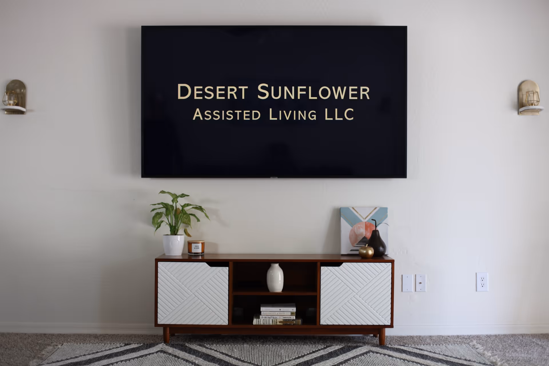 A modern living room setup with a wall-mounted flat screen TV displaying the text 'Desert Sunflower Assisted Living LLC'. Below the TV is a wooden console table with white textured cabinet doors, decorated with a potted plant, a candle, a small vase, a decorative painting, and some books. The room has a light-colored wall and carpeted floor with a patterned rug.