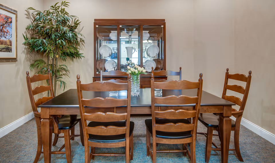 A dining room with a dark wooden table surrounded by six matching wooden chairs with black seats. A glass vase with white flowers is centered on the table. Behind the table is a wooden china cabinet displaying white dishes and glassware. To the left, there is a tall green potted plant and a framed picture on the beige wall.