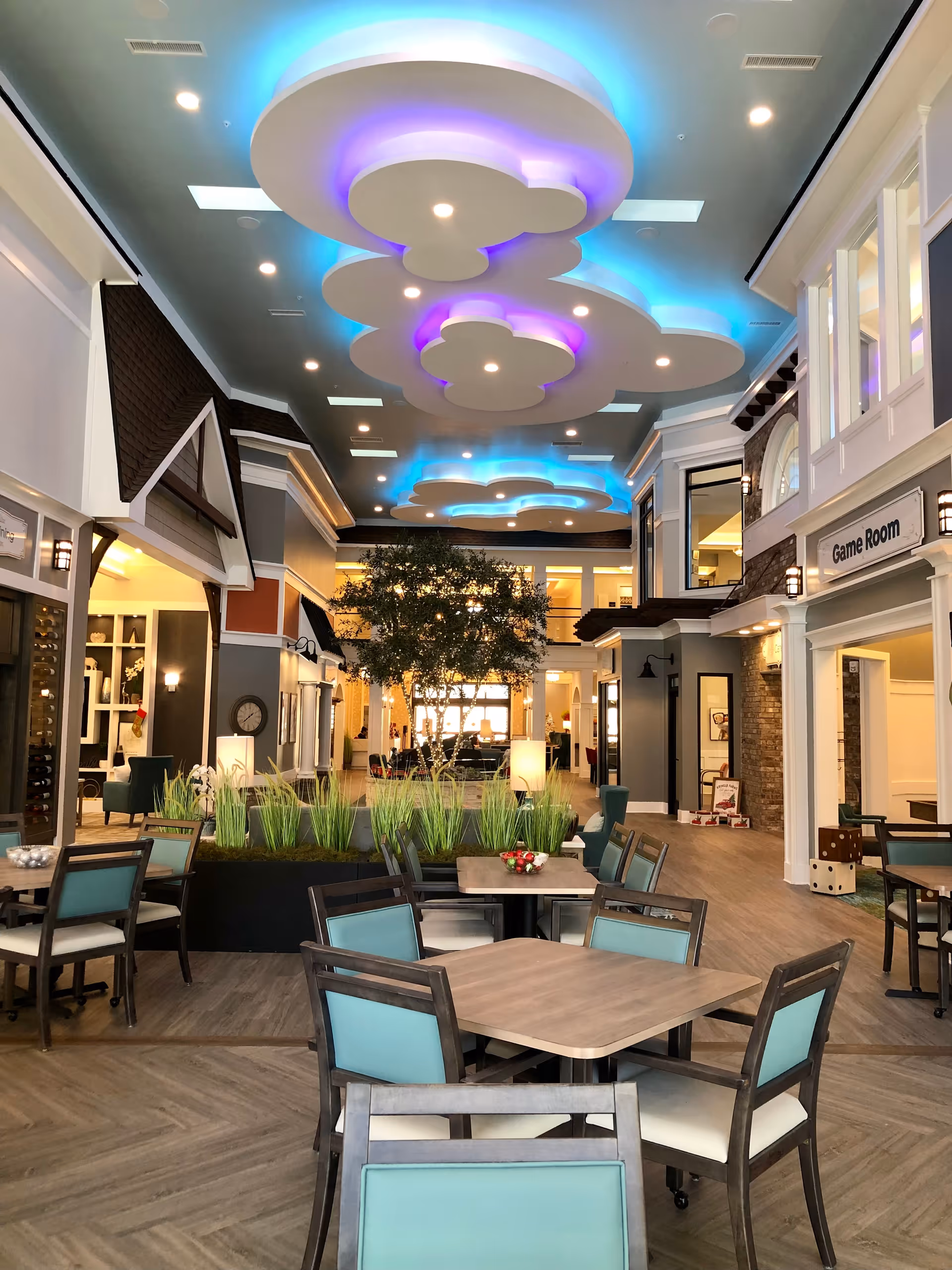 Interior view of a senior living facility common area with tables and chairs arranged for socializing or dining. The ceiling features decorative cloud-shaped light fixtures with blue and purple lighting. There are plants in the center and a sign indicating a game room on the right side.