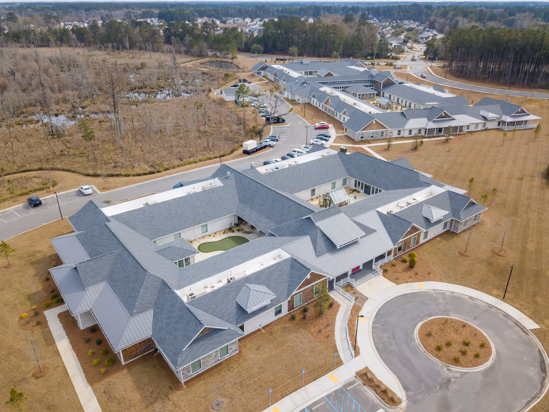 Aerial view of Legacy at Savannah Quarters senior living facility showing multiple connected buildings with gray roofs, parking lots with cars, surrounding grassy areas, and nearby wooded land.