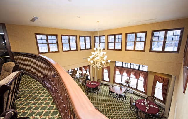 View from an upper balcony overlooking a spacious common area with multiple round tables covered with burgundy tablecloths and chairs. The room features large windows with brown curtains, a green patterned carpet, and a large chandelier hanging from the ceiling.