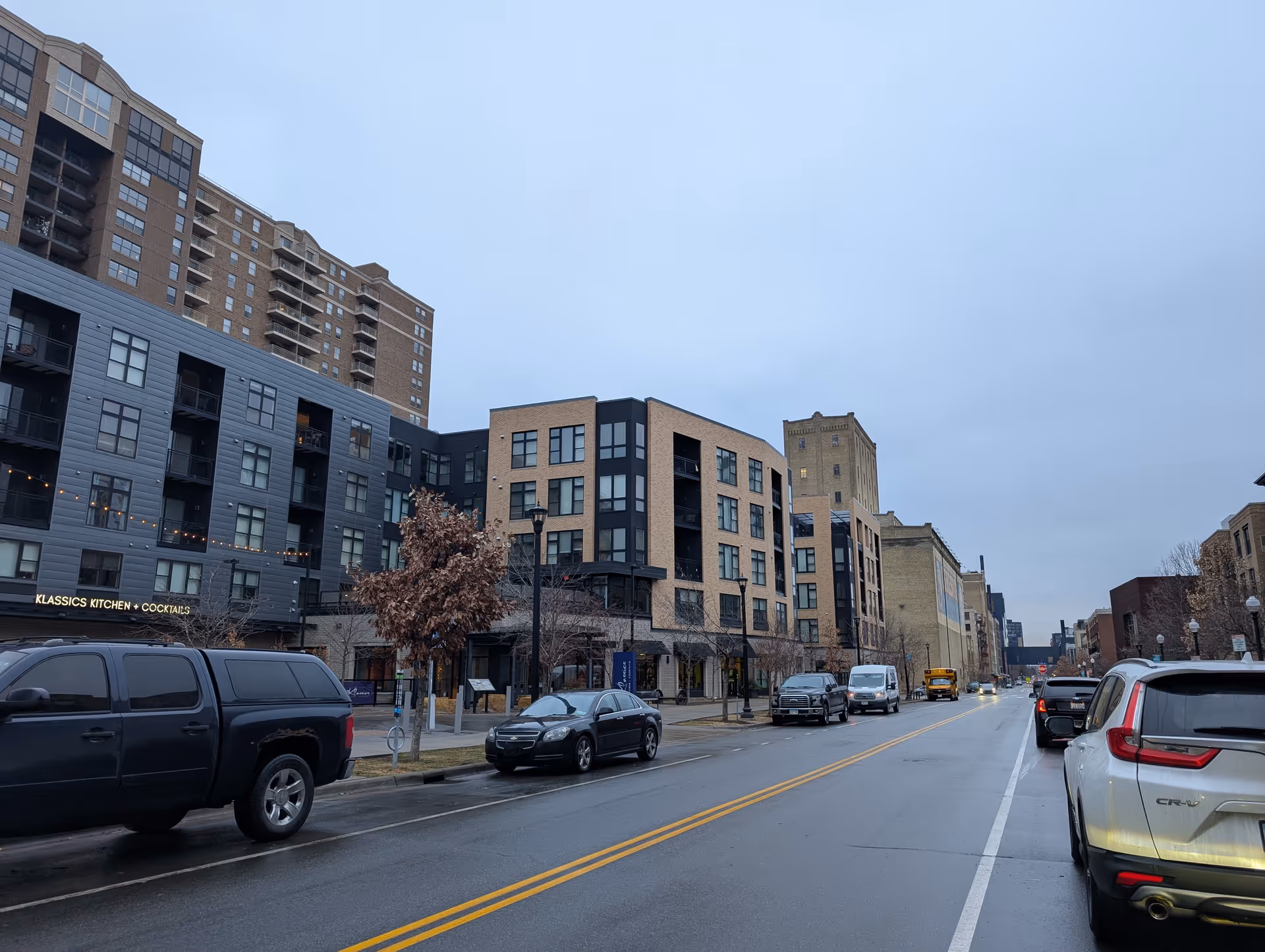 Street view of a city block with modern apartment buildings, parked cars, and a wet road on an overcast day.