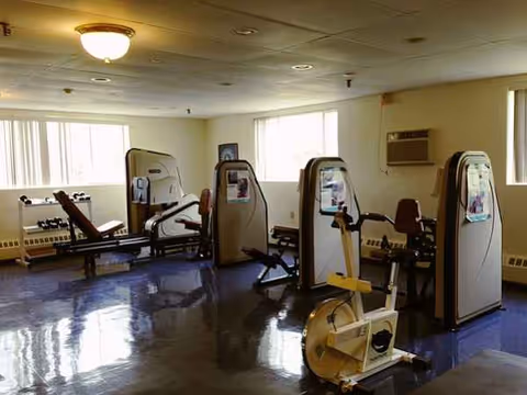 Indoor fitness center with multiple exercise machines and large windows at a senior facility.