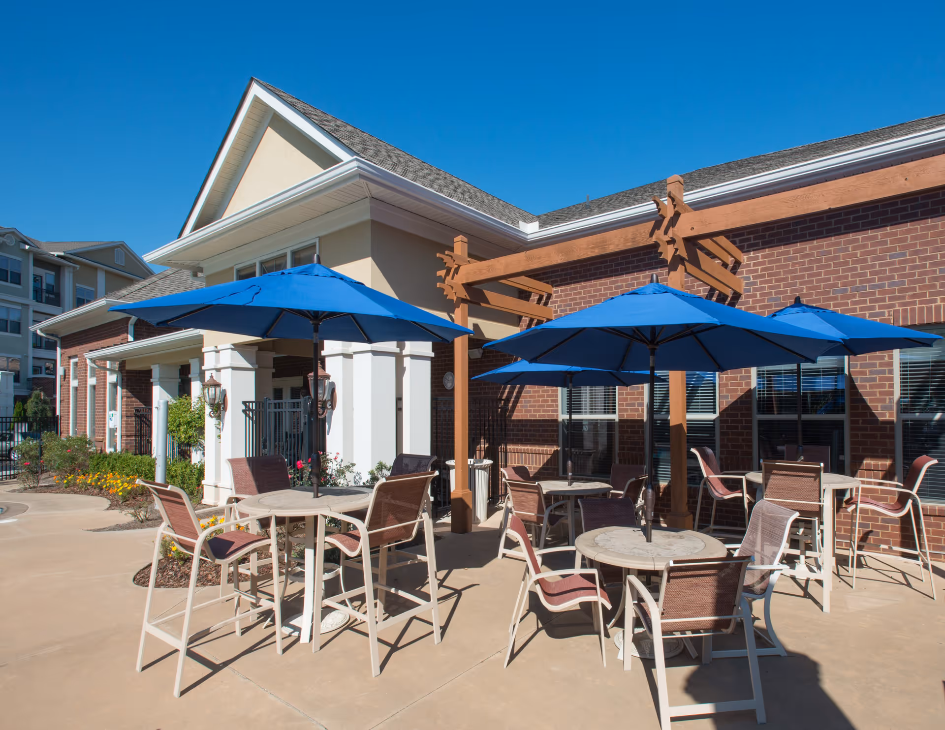 Outdoor patio area at Park at Johns Creek with round tables, chairs, and blue umbrellas providing shade. The patio is adjacent to a brick building with windows and a wooden pergola structure. The sky is clear and blue.