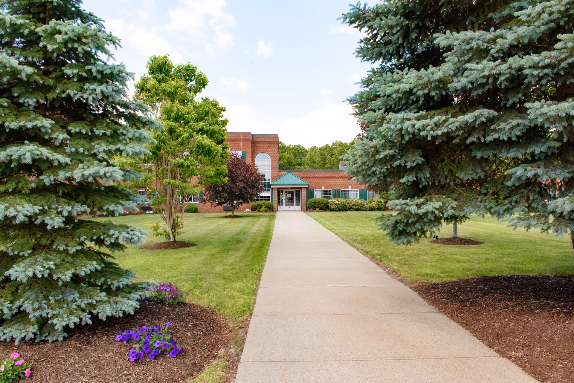A paved walkway leading to the entrance of a brick building with a green roof over the door, surrounded by well-maintained lawns, trees, and flower beds under a partly cloudy sky.