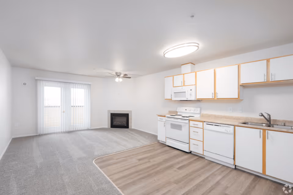 An open-concept living area and kitchen in a senior living facility. The kitchen features white cabinets with wood trim, a white stove, microwave, dishwasher, and sink. The living area has carpeted flooring, a ceiling fan with lights, a fireplace, and sliding glass doors with vertical blinds leading to an outdoor space.