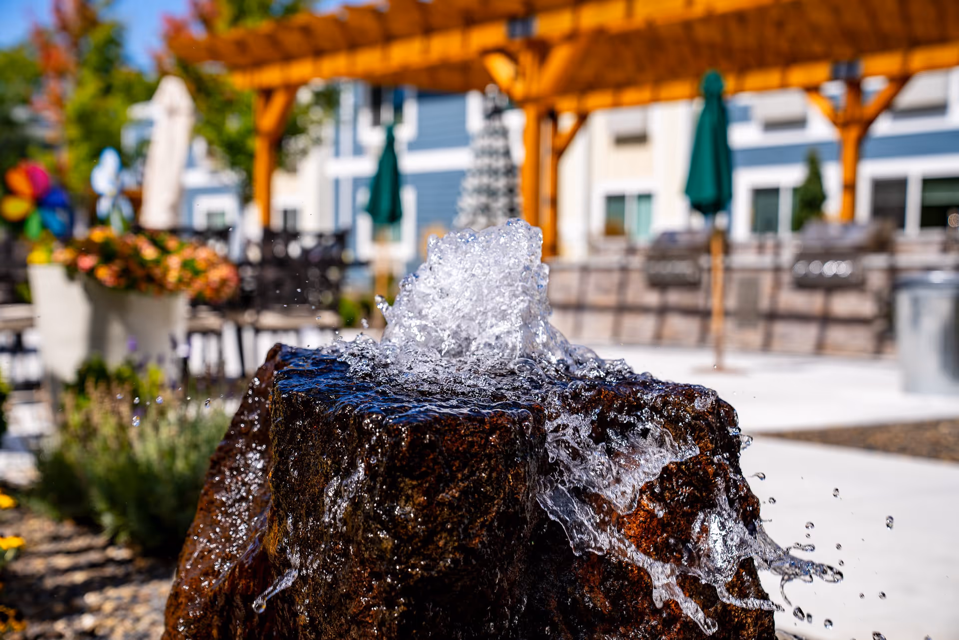 Close-up of a water fountain with water splashing over a dark stone in an outdoor patio area with tables, chairs, umbrellas, and a wooden pergola in the background.