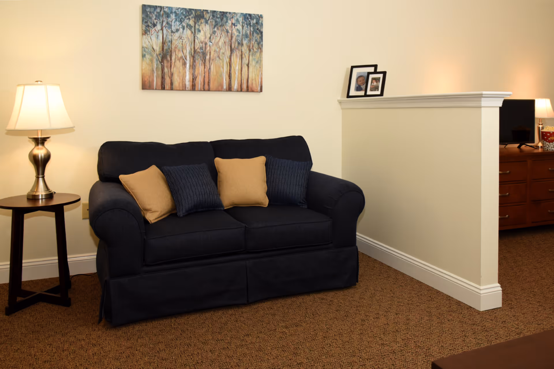A cozy living room area with a dark blue loveseat adorned with two tan and two dark blue pillows. To the left of the loveseat is a small round wooden side table with a lamp on it. Above the loveseat hangs a painting of trees with a soft, warm color palette. To the right, a half wall separates the living room from another area with a wooden dresser, a TV, and a lamp. The room has beige walls and brown carpeted flooring.