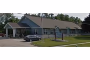 Single-story building with a gable roof and multiple windows, surrounded by a well-maintained lawn and a parking area with a car parked. A flagpole with an American flag is in front of the building, along with a sign indicating the facility name.