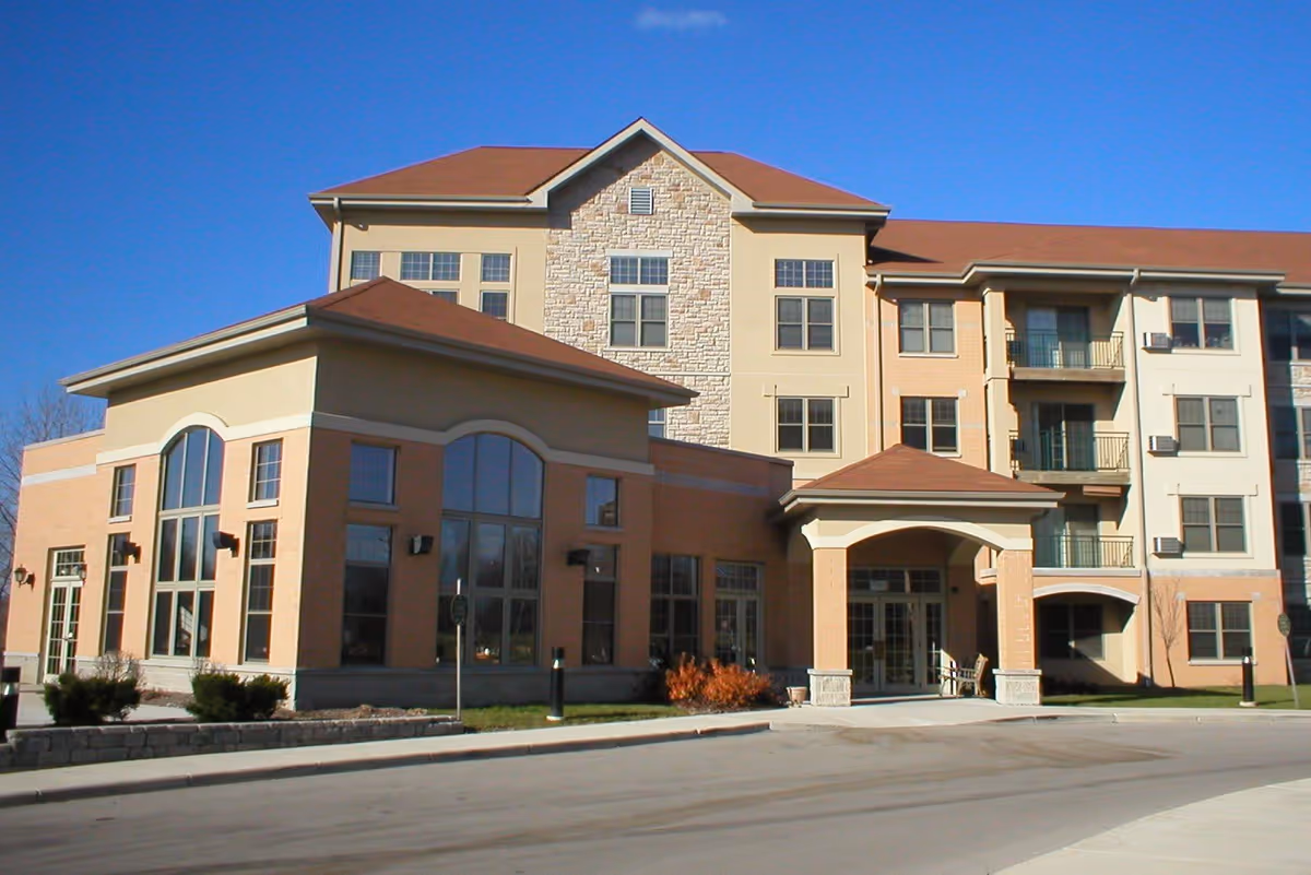 Exterior view of a multi-story assisted living facility building with large windows, a covered entrance, and a clear blue sky.