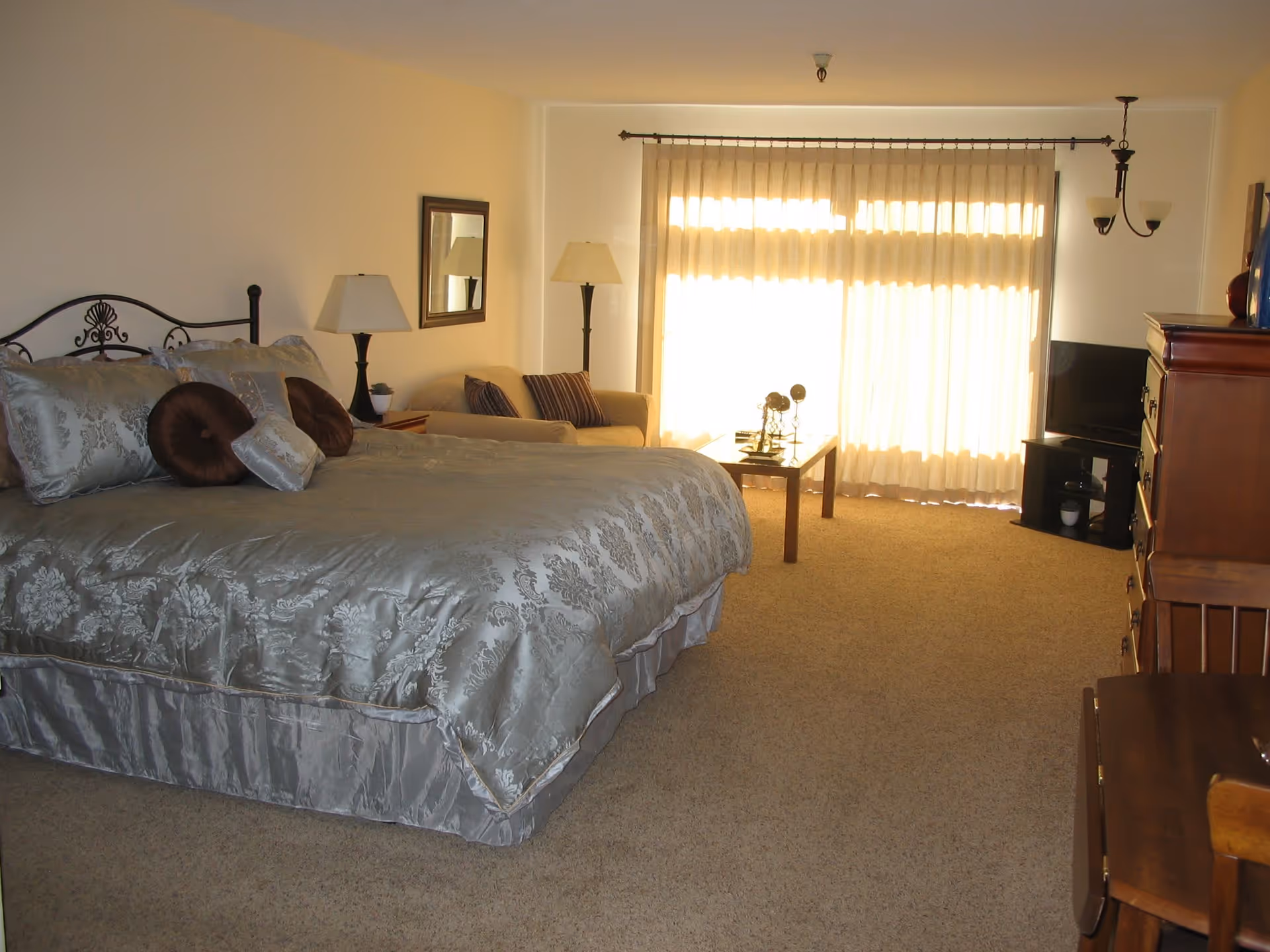 A spacious bedroom with a large bed covered in silver patterned bedding and multiple pillows. There is a beige sofa with striped cushions, a wooden coffee table with decorative items, a floor lamp, a table lamp on a nightstand, a wall mirror, a wooden dresser, a small TV on a stand, and large windows with sheer curtains letting in natural light.