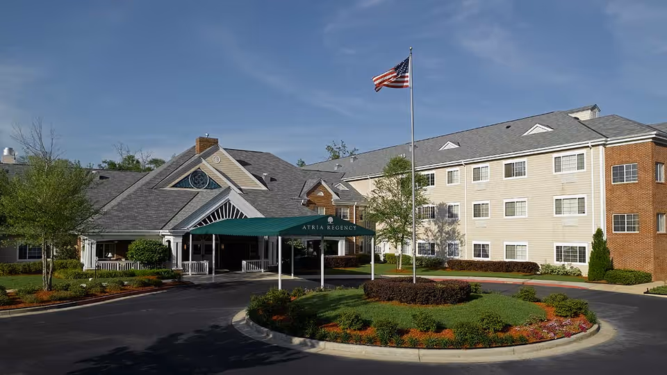 Exterior view of a senior living facility with a circular driveway and landscaped roundabout featuring an American flag on a flagpole. The building is three stories tall with beige siding and brick accents, and a covered entrance with a green awning labeled 'ATRIA REGENCY'.