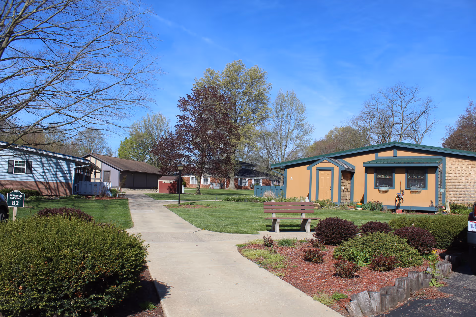 A sunny outdoor scene at a senior living facility showing a paved walkway leading between small residential buildings surrounded by green lawns, bushes, and trees with a clear blue sky overhead. There is a wooden bench along the walkway and a reserved parking sign labeled B2 on the left side.