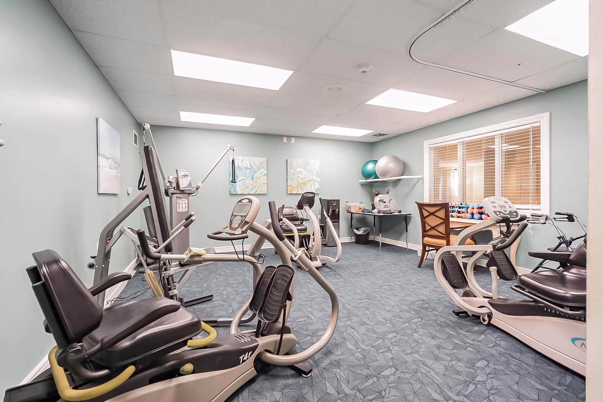 A small fitness room with exercise equipment including stationary bikes and a multi-function weight machine. The room has light blue walls, a blue patterned carpet, fluorescent ceiling lights, and two framed artworks on the wall. There is a table with exercise balls and dumbbells near a window with blinds, and a wooden chair is placed next to the table.