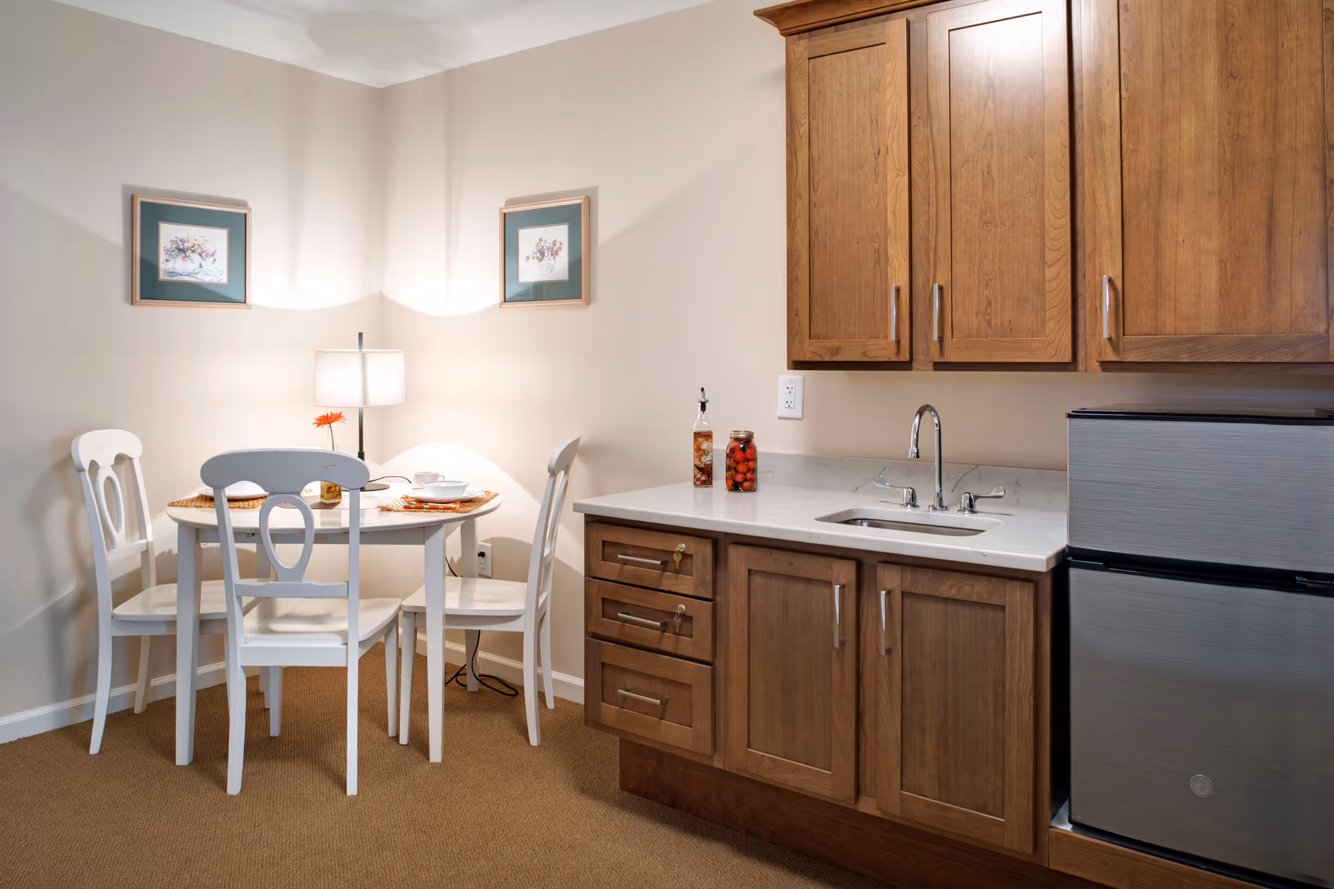 A small dining area with a round white table and four white chairs. The table is set with placemats, bowls, and a small flower vase. Next to the dining area is a kitchenette with wooden cabinets, a countertop with a sink, and a small stainless steel refrigerator. Two framed floral pictures hang on the beige walls, and a table lamp provides soft lighting.