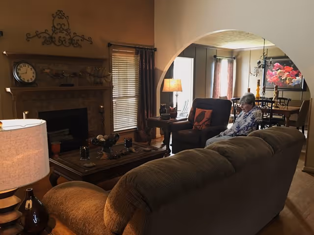 Cozy living room with a sofa, coffee table, fireplace, and an elderly woman seated in a chair near an archway to the dining area.